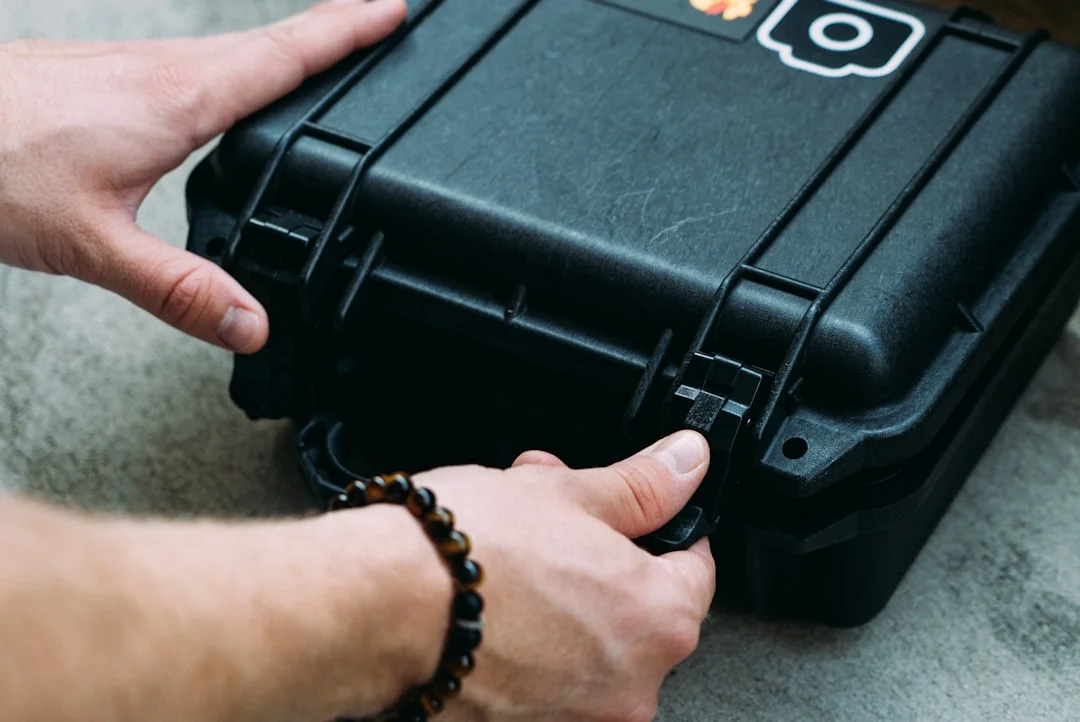 A broadcast cinematographer loading a Pelican 1510 carry-on hard case with camera gear in an airport terminal, overhead bins visible in the background